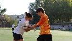 Dos jugadoras de fútbol ayudándose una a otra. Una de ellas le pasa el brazadete de capitán del equipo a la otra.